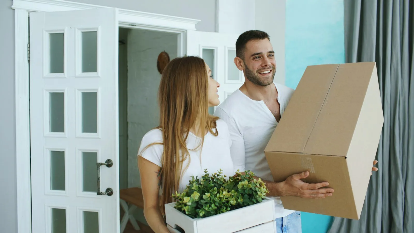 A smiling couple carrying a moving box and a potted plant through the front door of their new home.